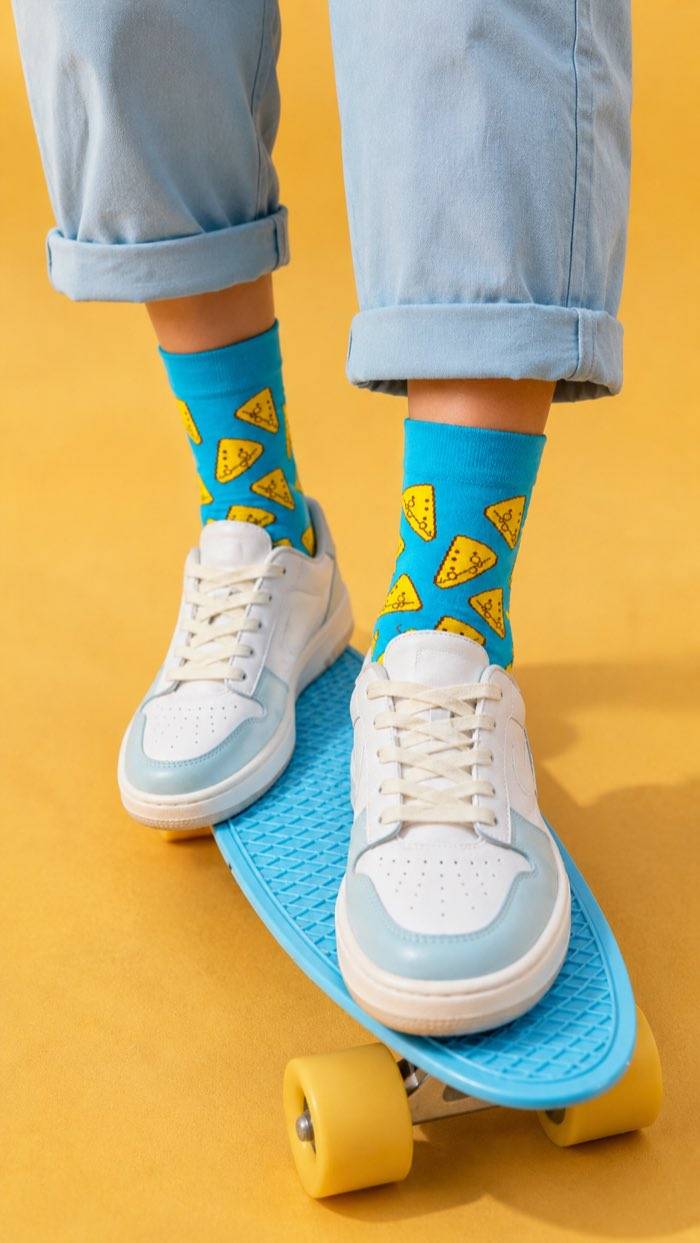 Blue socks with yellow cheese slices, worn with white sneakers on a blue skateboard against a yellow background